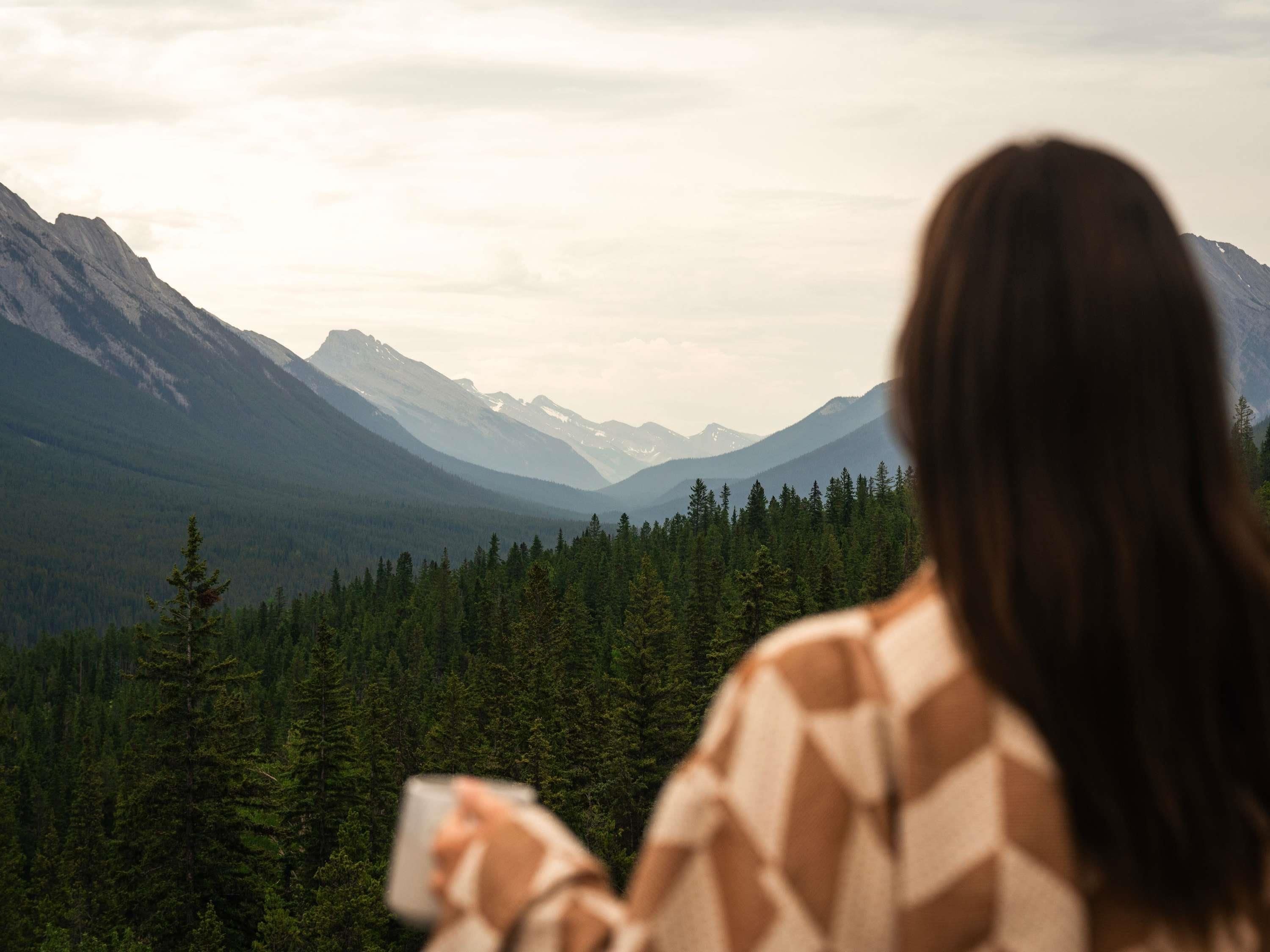 Una persona de cabello largo sosteniendo una taza, con vistas a un paisaje montañoso con bosques densos.