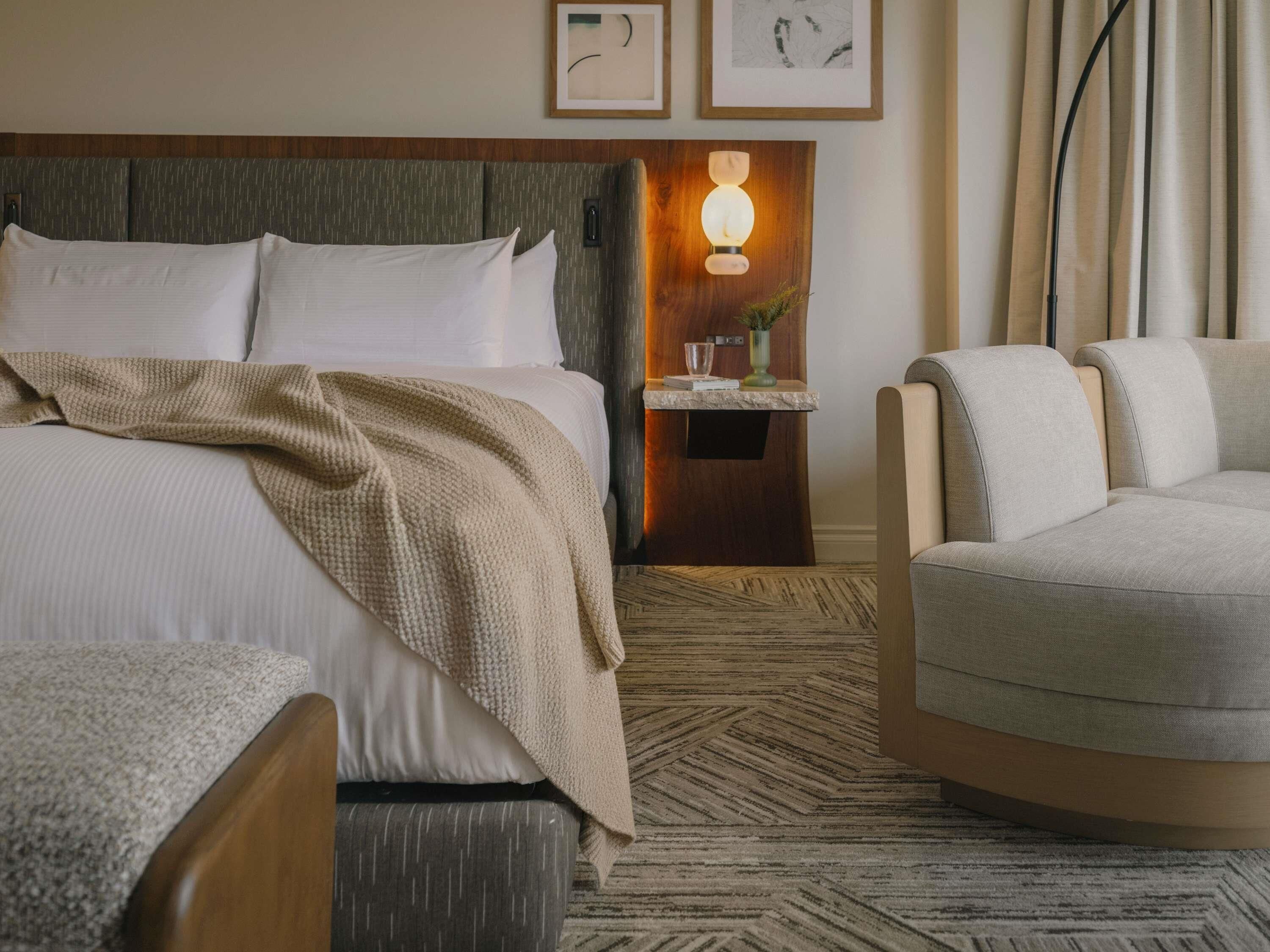 A hotel room with a large bed, a bedside table with a lamp, a chair, and a patterned carpet.