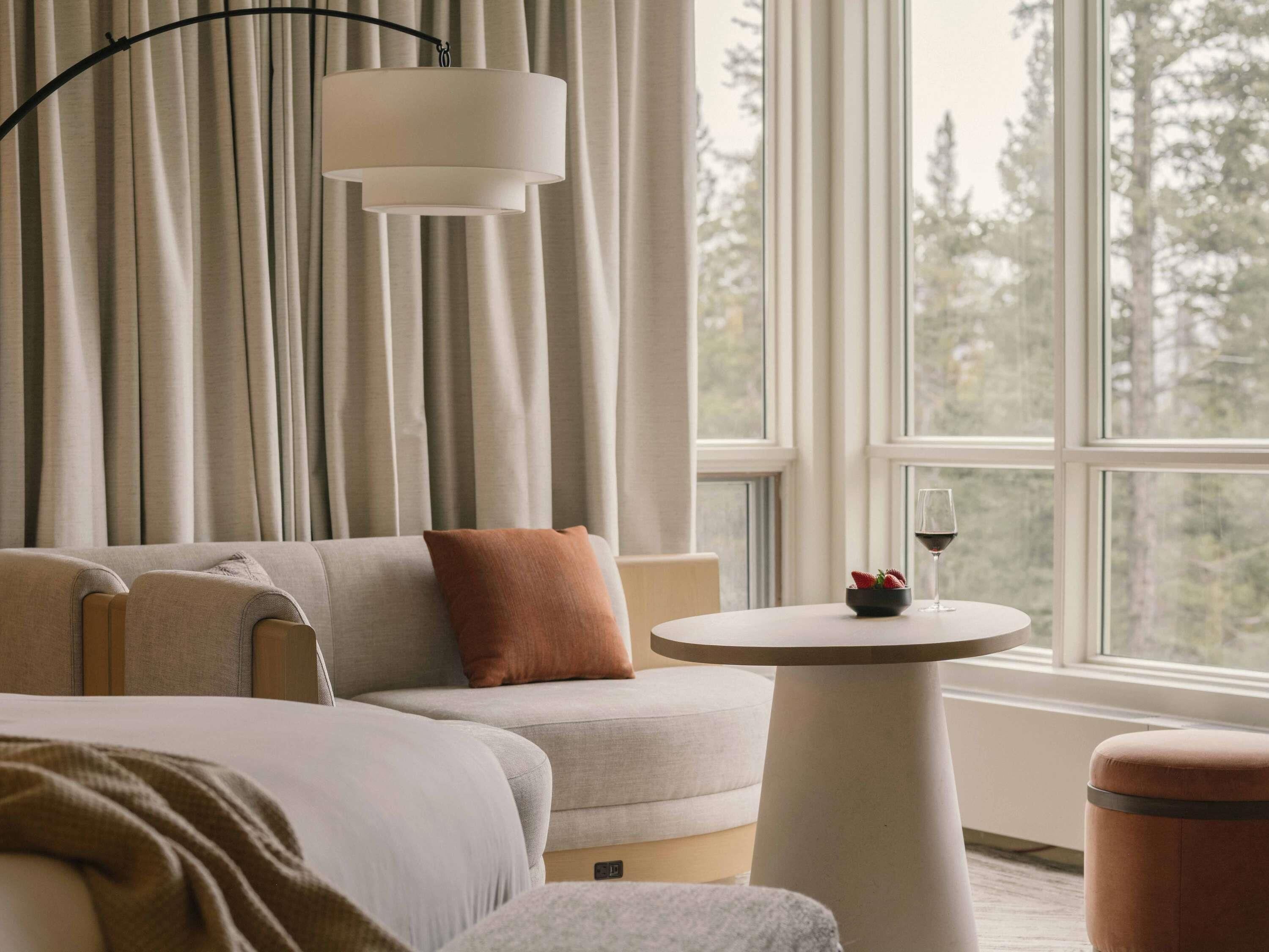A modern hotel room with a sofa, a round table, and a view of trees through the window.