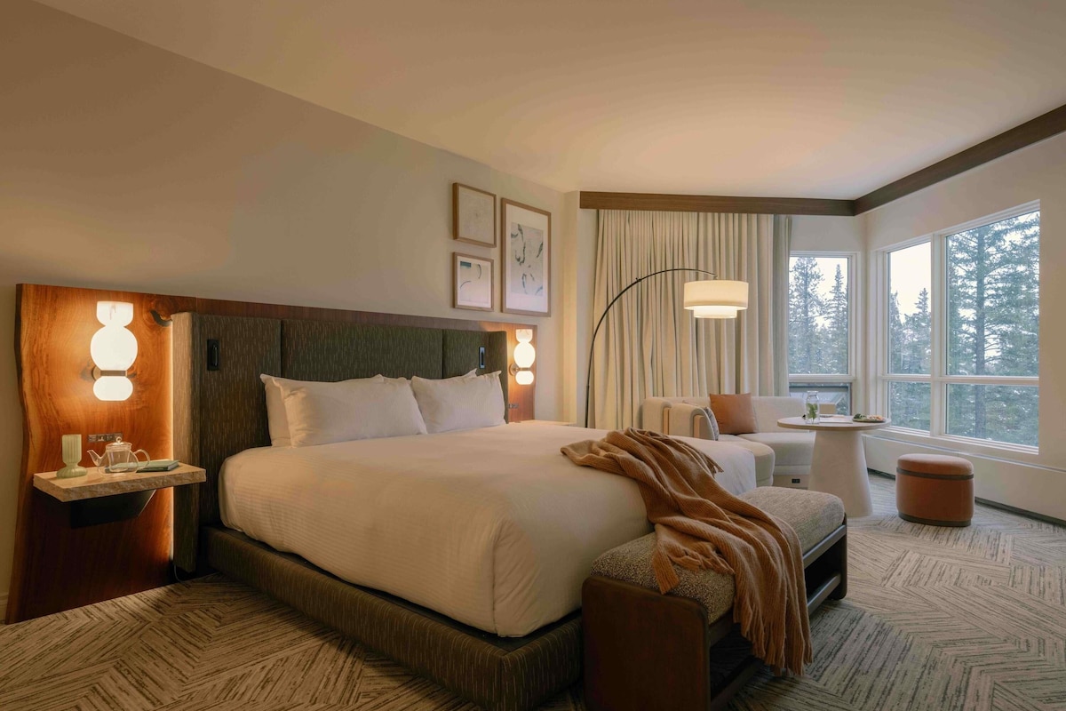 Una habitación de hotel con una cama grande, una mesita de noche con lámpara, una silla, una pequeña mesa con lámpara y una ventana con vistas a árboles.