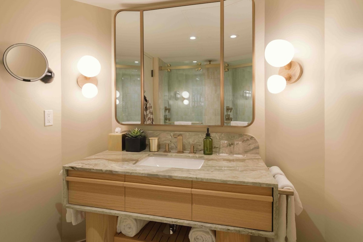 Un baño con un espejo grande, dos luces empotradas, un lavamanos y un mueble de tocador de madera con cajones y un perchero para toallas.