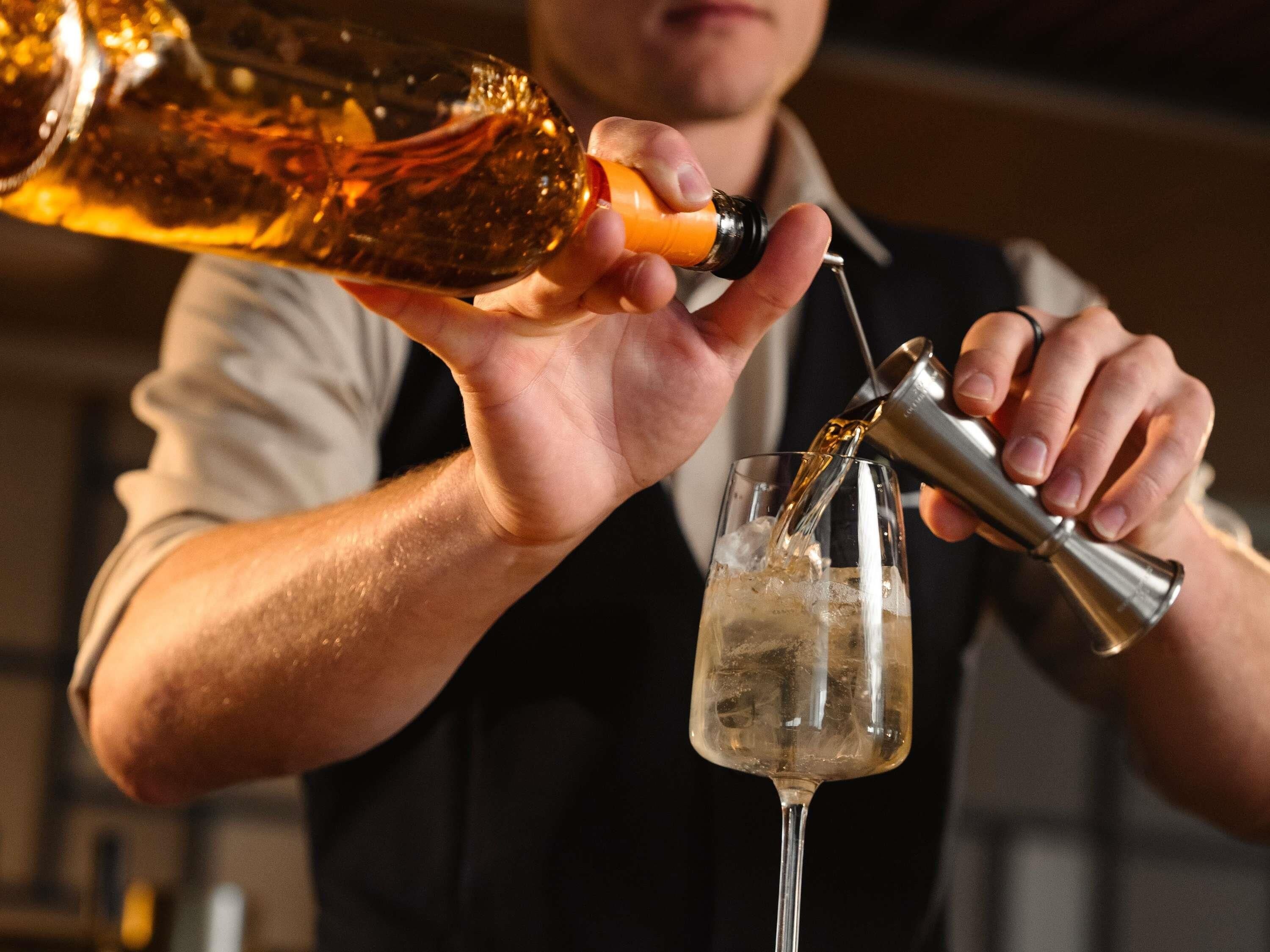 A person pouring a liquid from a bottle into a glass using a jigger.