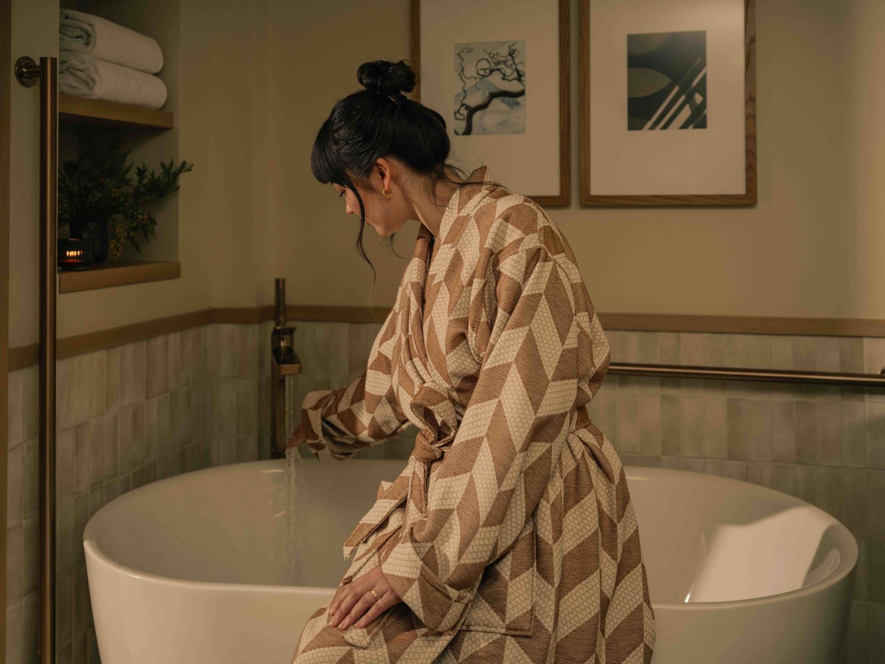 A person in a patterned bathrobe sitting in a bathtub with a towel rack above and framed artwork on the wall.