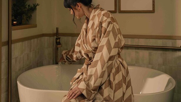 Una persona en un albornoz estampado sentada en una bañera, con un perchero arriba y una obra de arte en la pared.