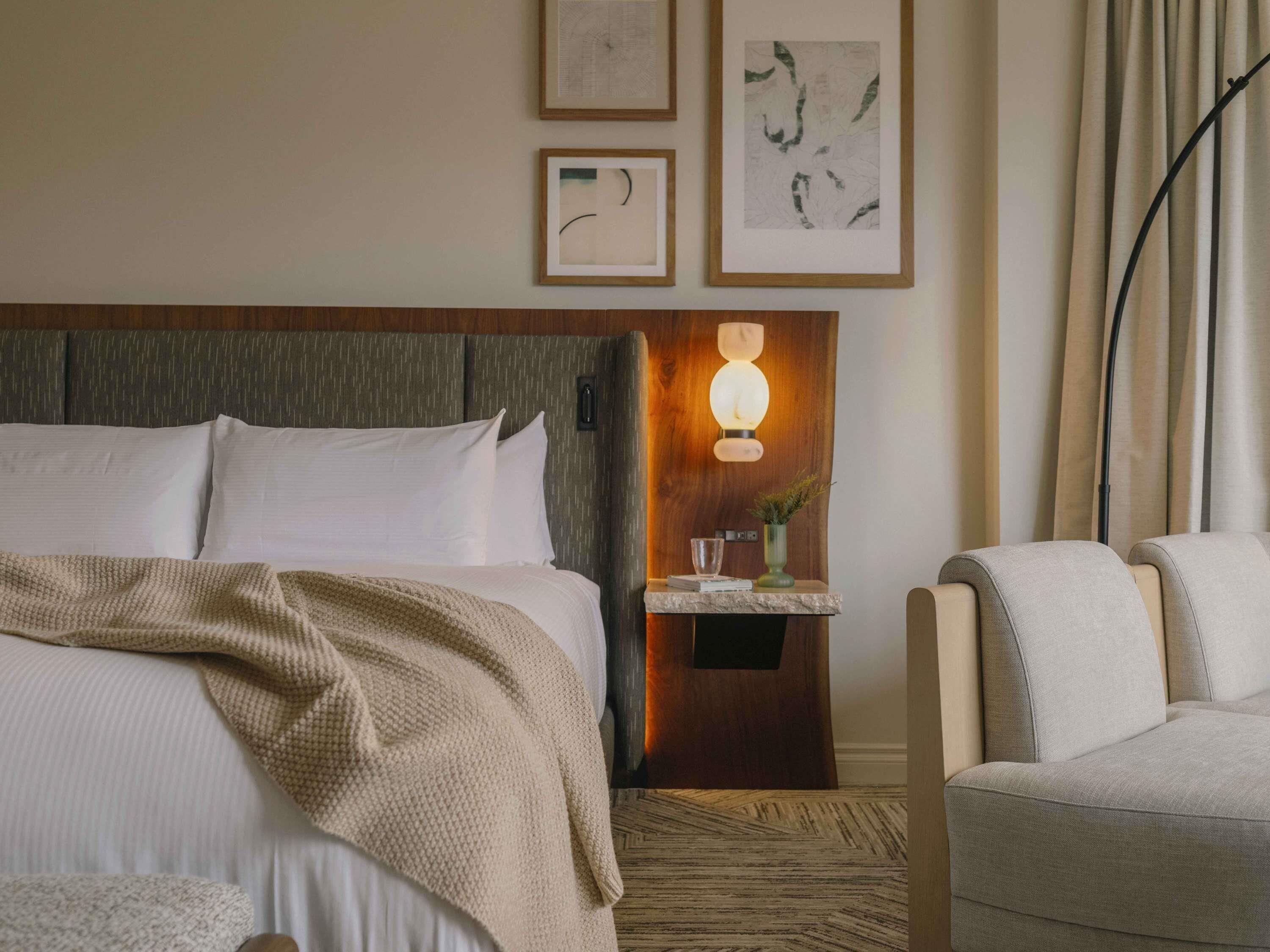 Una camera d'albergo con un letto, un comodino con una lampada, una sedia e un'opera d'arte appesa al muro.
