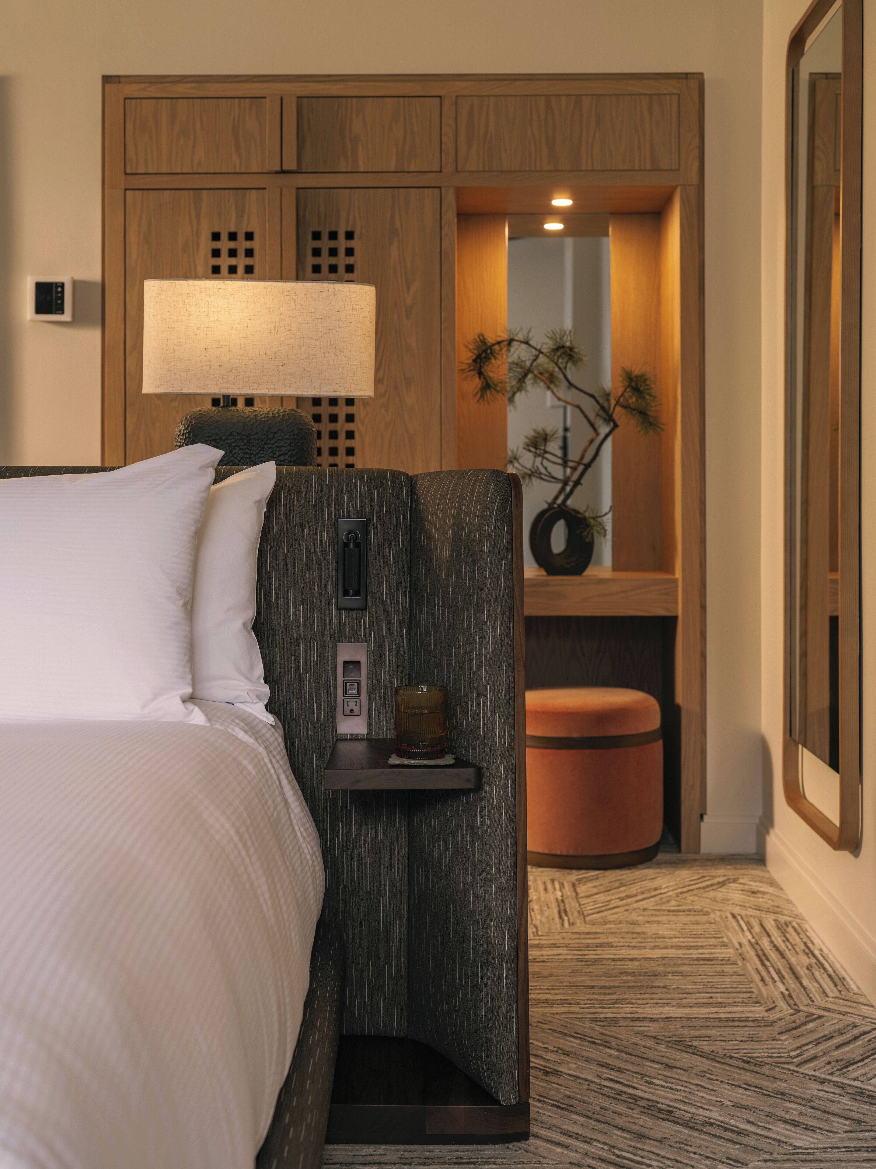 A hotel room with a bed, a nightstand, a lamp, a small table with a glass, and a mirror.