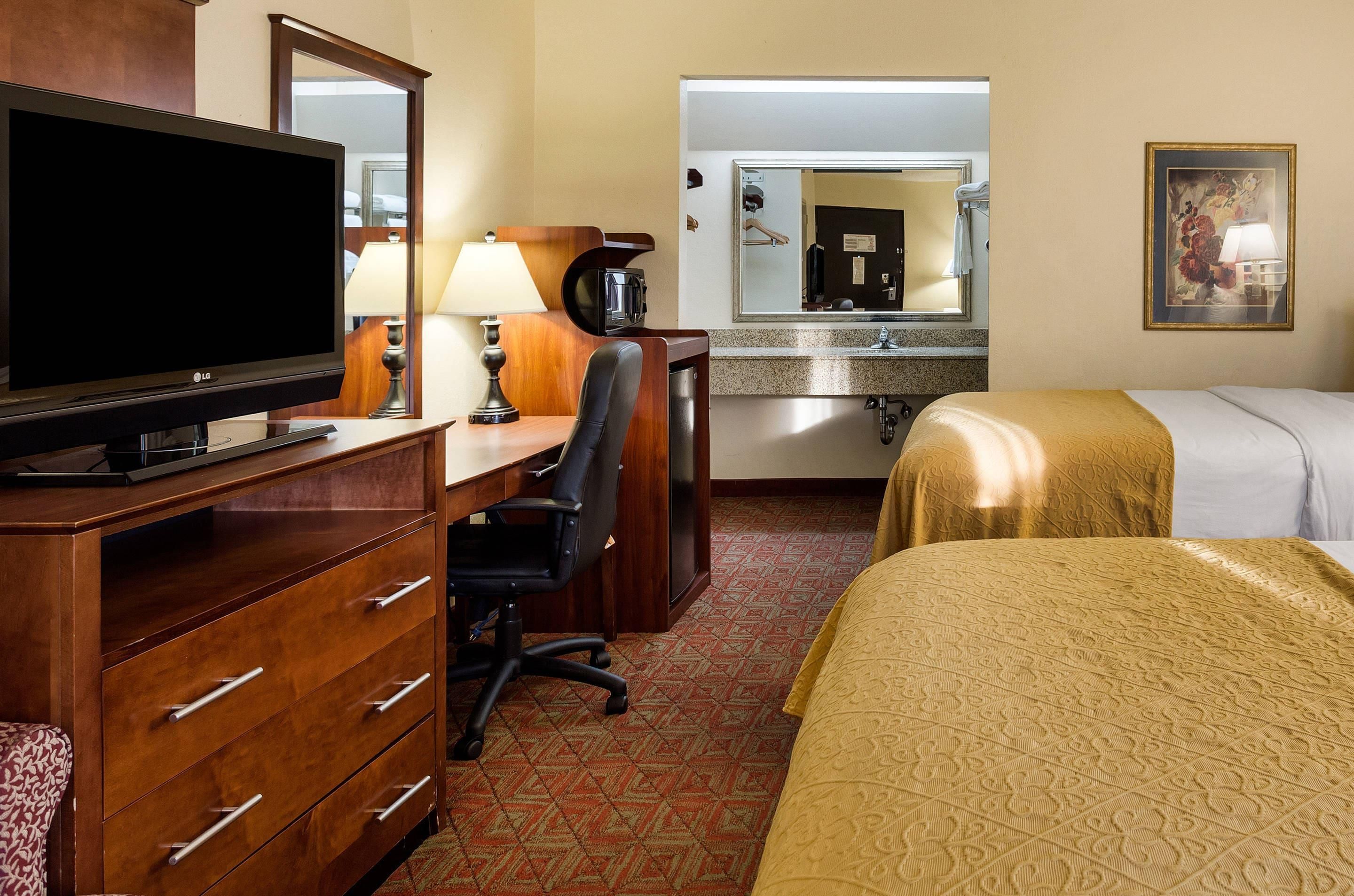 Quarto de hotel com televisão, escrivaninha, cadeira, cama e espelho de banheiro.