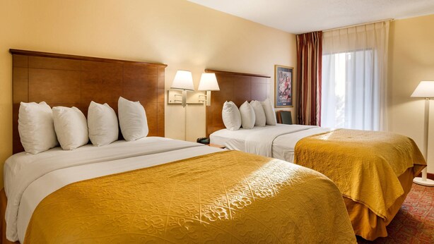 Hotelkamer met twee bedden, elk voorzien van een hoofdbord, wit beddengoed en een gele accentdeken. Twee bedlampen, een raam met gordijnen en een ingelijste foto aan de muur.