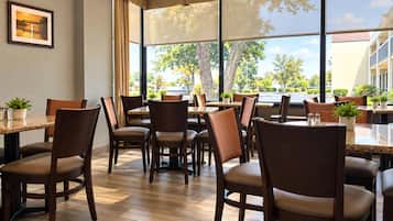 A restaurant with multiple tables and chairs, large windows with a view of trees and buildings, and a framed picture on the wall.