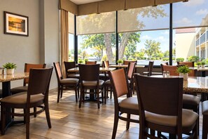 A restaurant with multiple tables and chairs, large windows with a view of trees and buildings, and a framed picture on the wall.