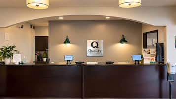A hotel reception desk with a sign reading 'Quality by Choice Hotels,' two wall-mounted lamps, a reception counter, a computer monitor, and a plant.