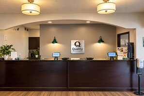 Ein Hotelrezeptionstresen mit einem Schild, auf dem 'Quality by Choice Hotels' steht, zwei Wandleuchten, eine Rezeptionstheke, ein Computermonitor und eine Pflanze.