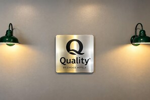 Ein an der Wand befestigtes Schild mit dem 'Quality'-Logo und dem Text 'by Choice Hotels', das von zwei grün beleuchteten Wandlampen eingerahmt wird.