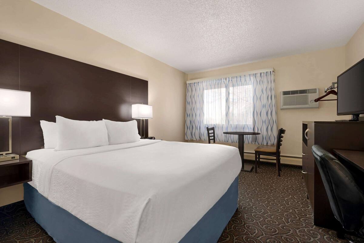 A hotel room with a large bed, bedside tables with lamps, a desk with a chair, a TV, and an air conditioning unit.