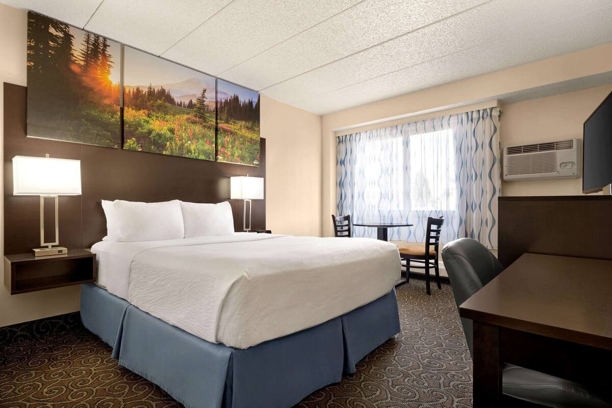 A hotel room with a large bed, two bedside tables with lamps, a desk with chairs, and a large window with curtains.
