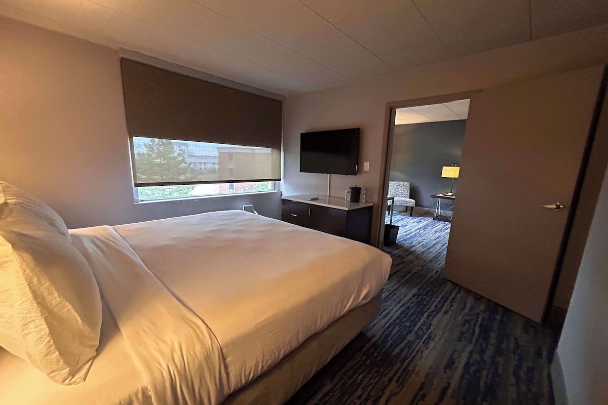 A hotel room with a large bed, a flat-screen TV mounted on the wall, a desk with a chair, and a small table with a lamp.