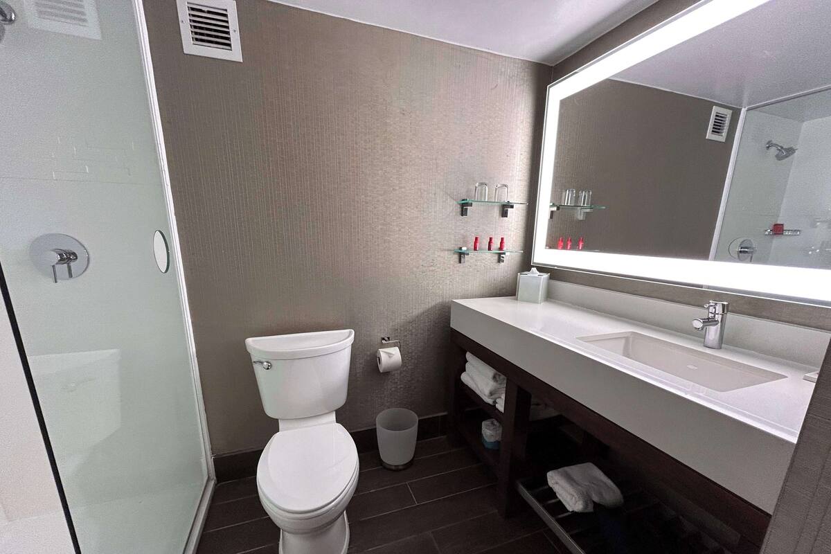 A modern bathroom with a toilet, sink, and mirror. There are toiletries on a shelf and a trash bin.