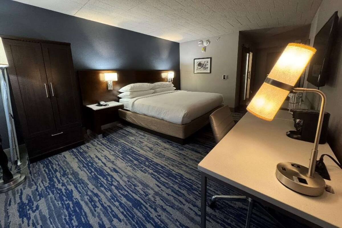 A hotel room with a bed, a desk, a chair, a wardrobe, and a lamp.