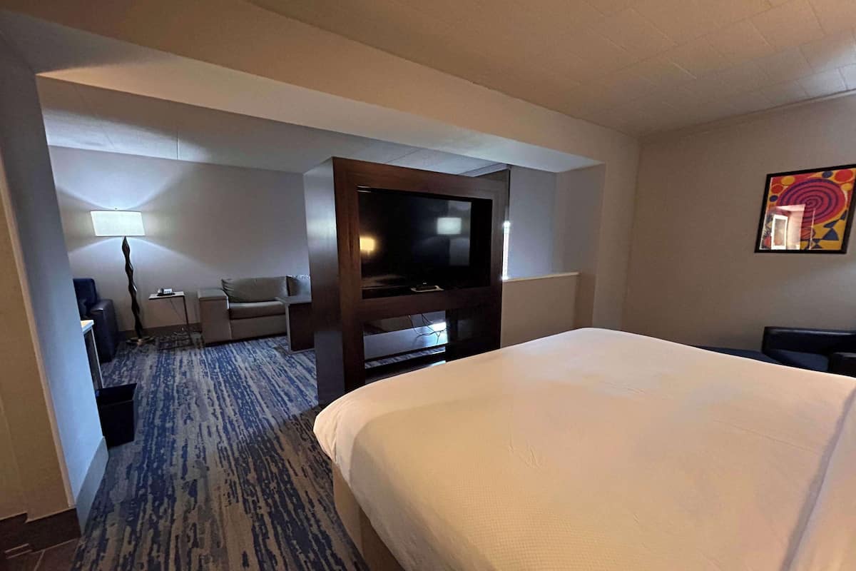 A hotel room with a large bed, a flat-screen TV mounted on a wooden stand, a sofa, a small table, and a lamp.
