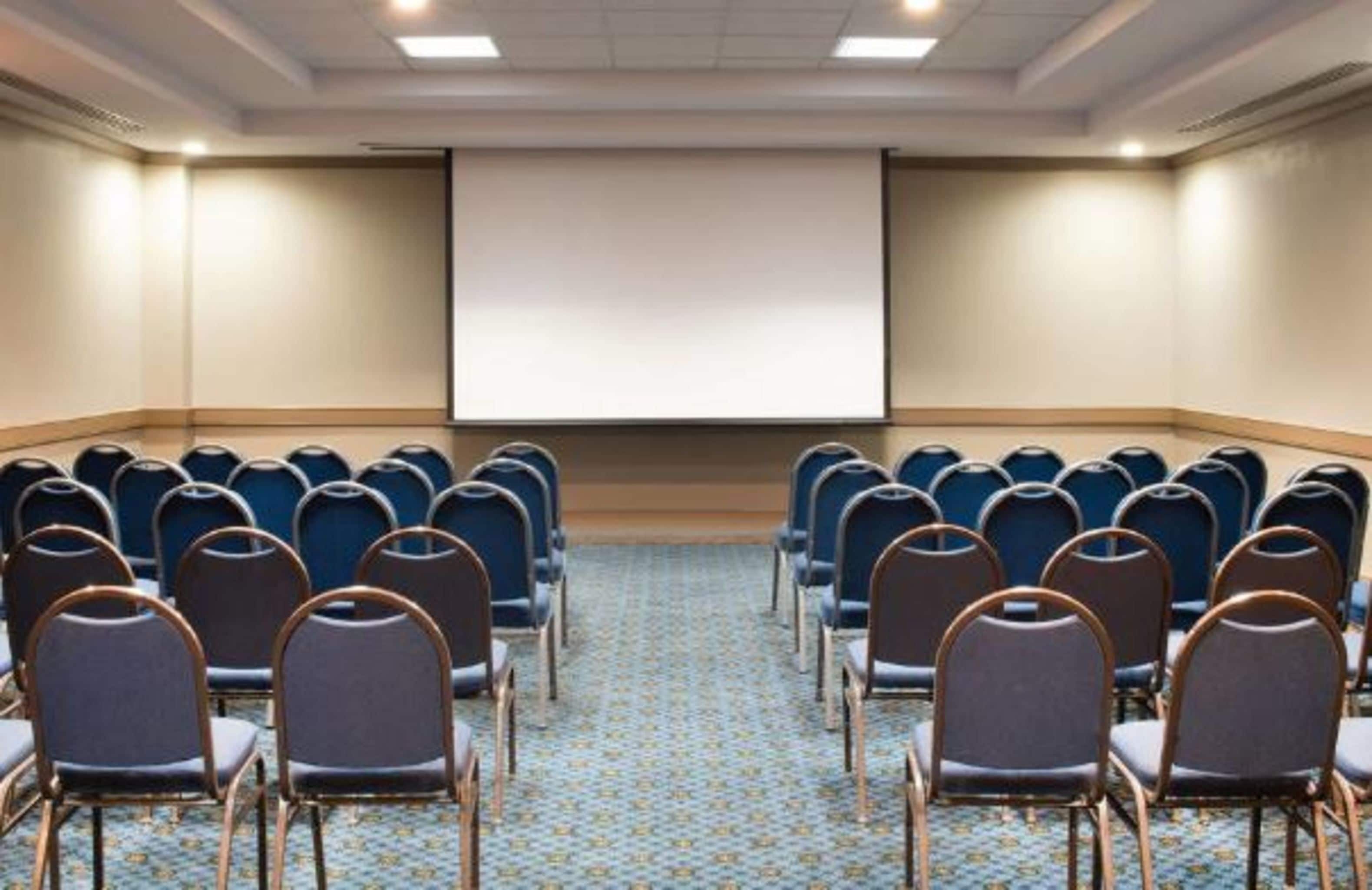 Un salón de conferencias con filas de sillas mirando hacia una pantalla de proyección.