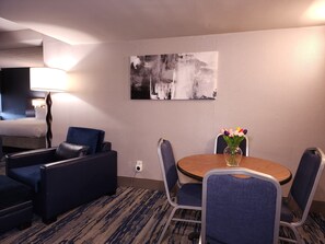 A hotel room with a bed, a desk, a chair, a table with a vase of flowers, and a painting on the wall.
