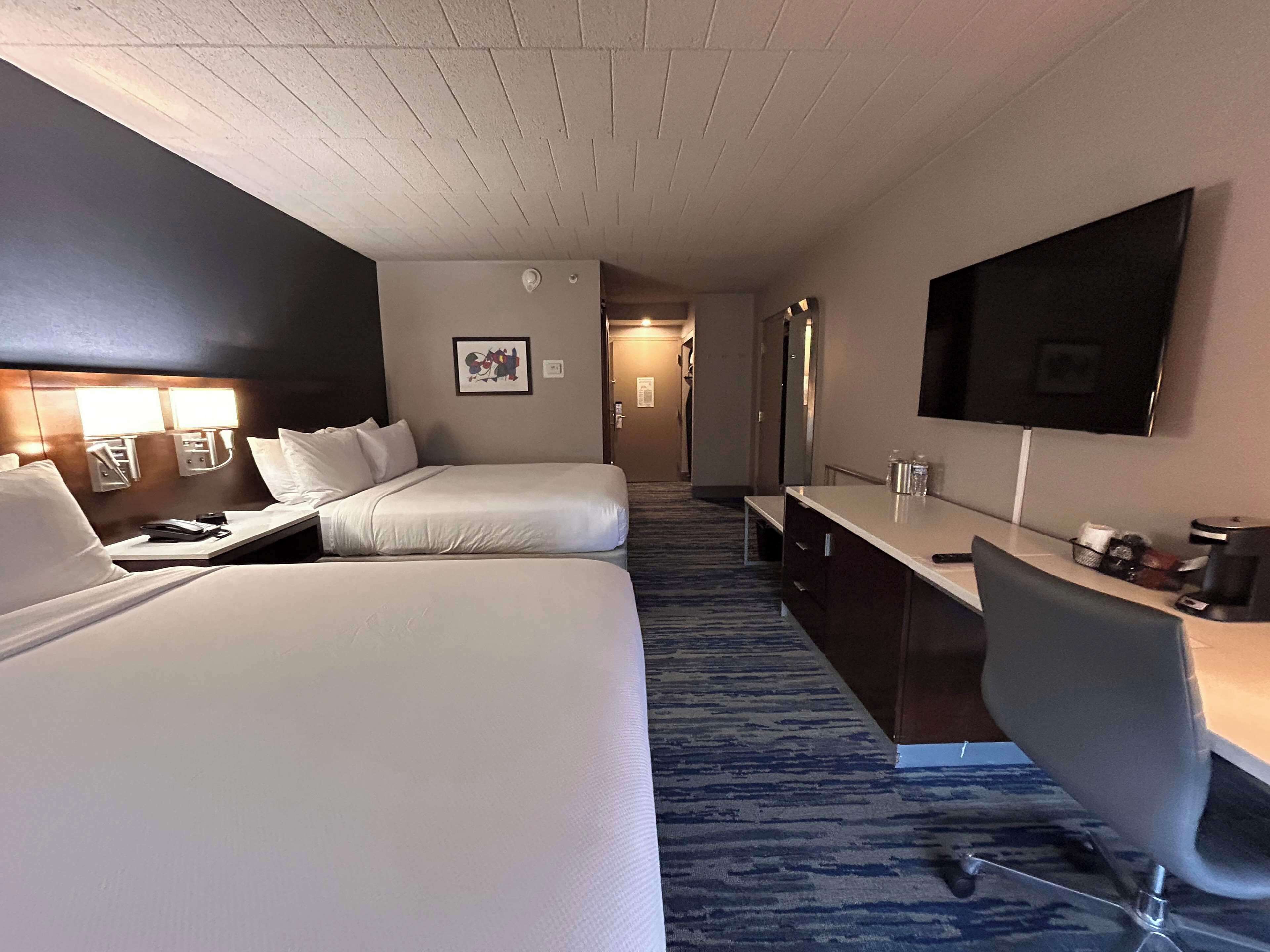 A hotel room with two beds, a desk, a chair, a TV, and a bathroom visible in the background.