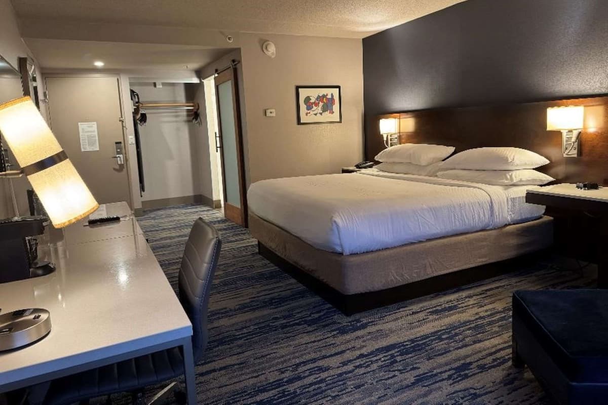 A hotel room with a large bed, a desk, and a chair. There is a door leading to a bathroom and a coat rack.