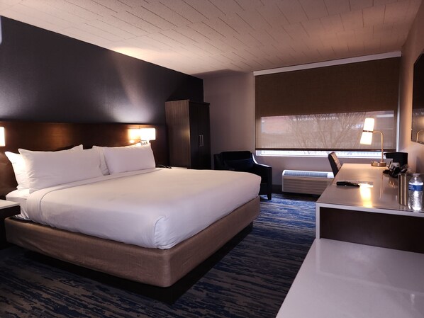 A hotel room with a large bed, a desk, a chair, and a window with blinds.