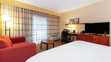 A hotel room with a large bed, a red sofa, a desk with a computer, a flat-screen TV, and a window with curtains.