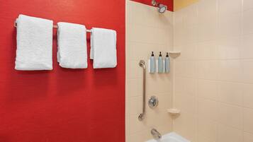 Un baño con una pared roja, tres toallas blancas colgadas en un perchero, una zona de ducha con puerta de cristal y un lavabo con artículos de aseo.
