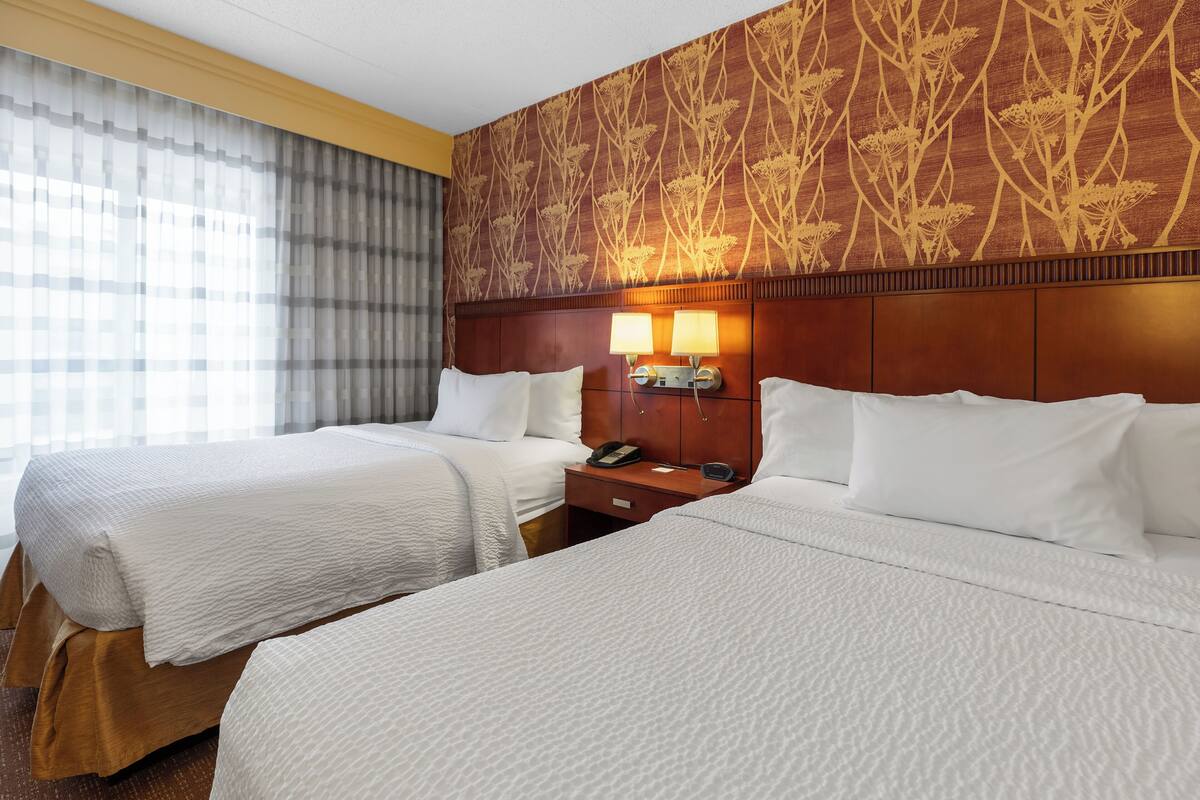 Habitación de hotel con dos camas, cabecera de madera, mesita de noche con lámpara y pared con estampado.