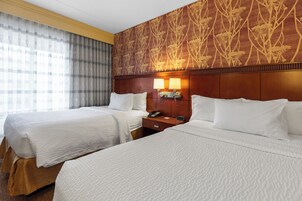Habitación de hotel con dos camas, cabecera de madera, mesita de noche con lámpara y pared con estampado.