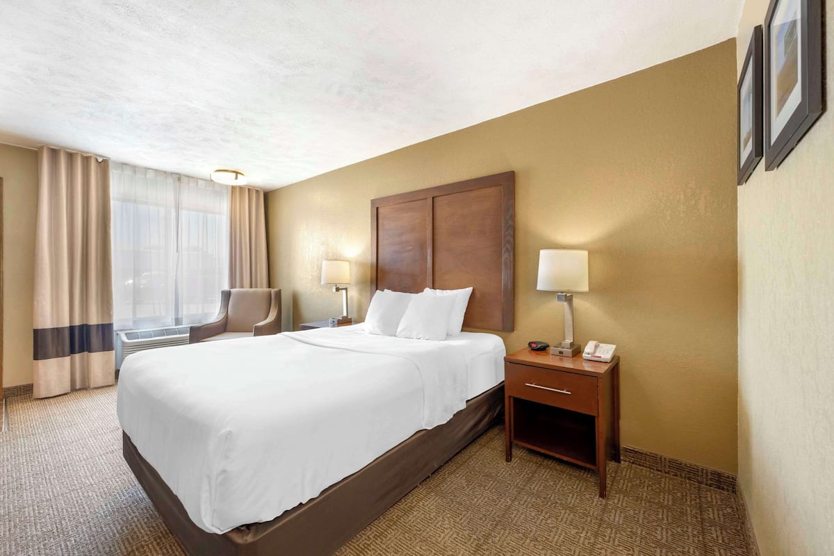 A hotel room with a large bed, a nightstand with a lamp, a chair, and a desk with a phone.