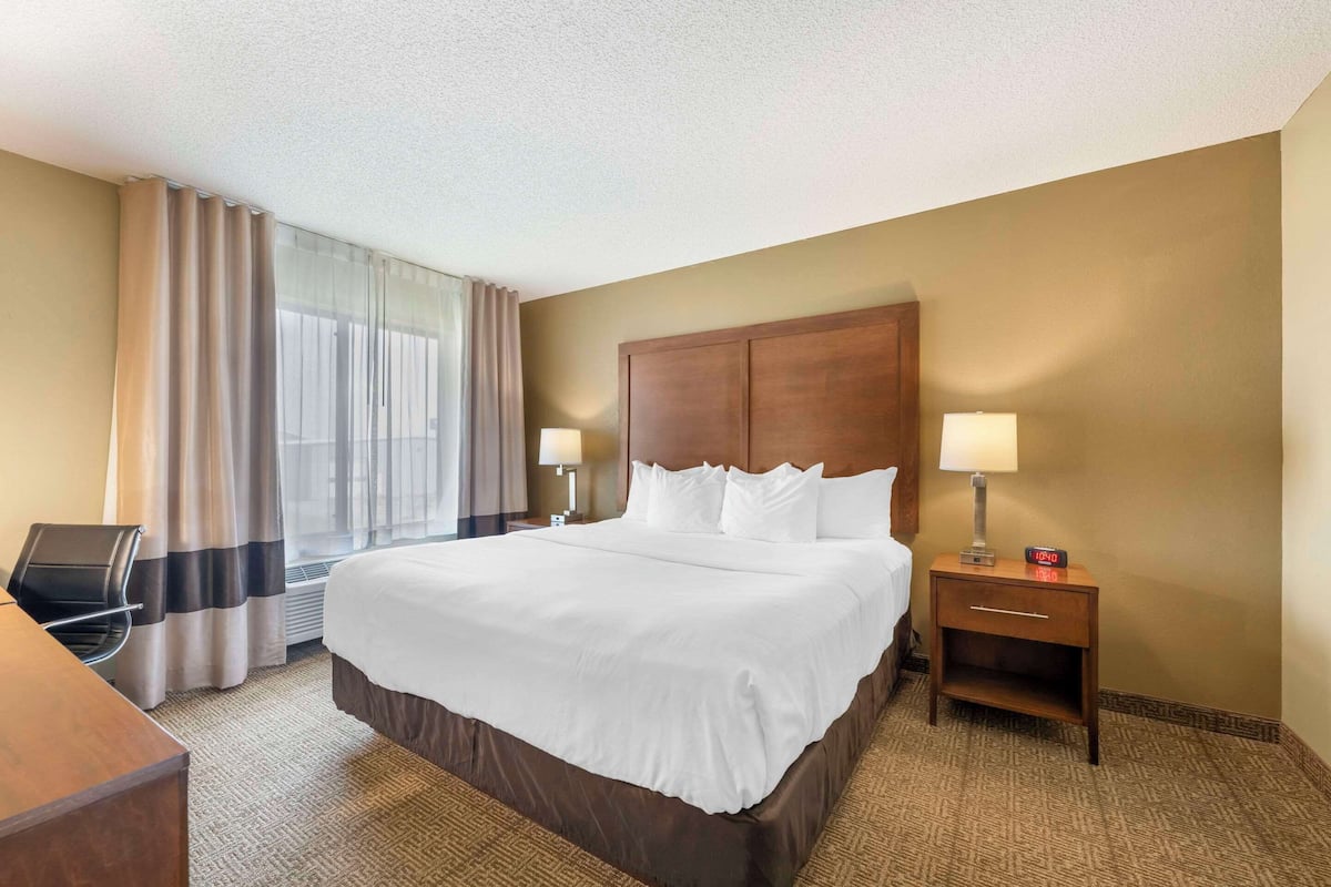 A hotel room with a large bed, two bedside lamps, a wooden nightstand, a desk with a chair, and a window with curtains.