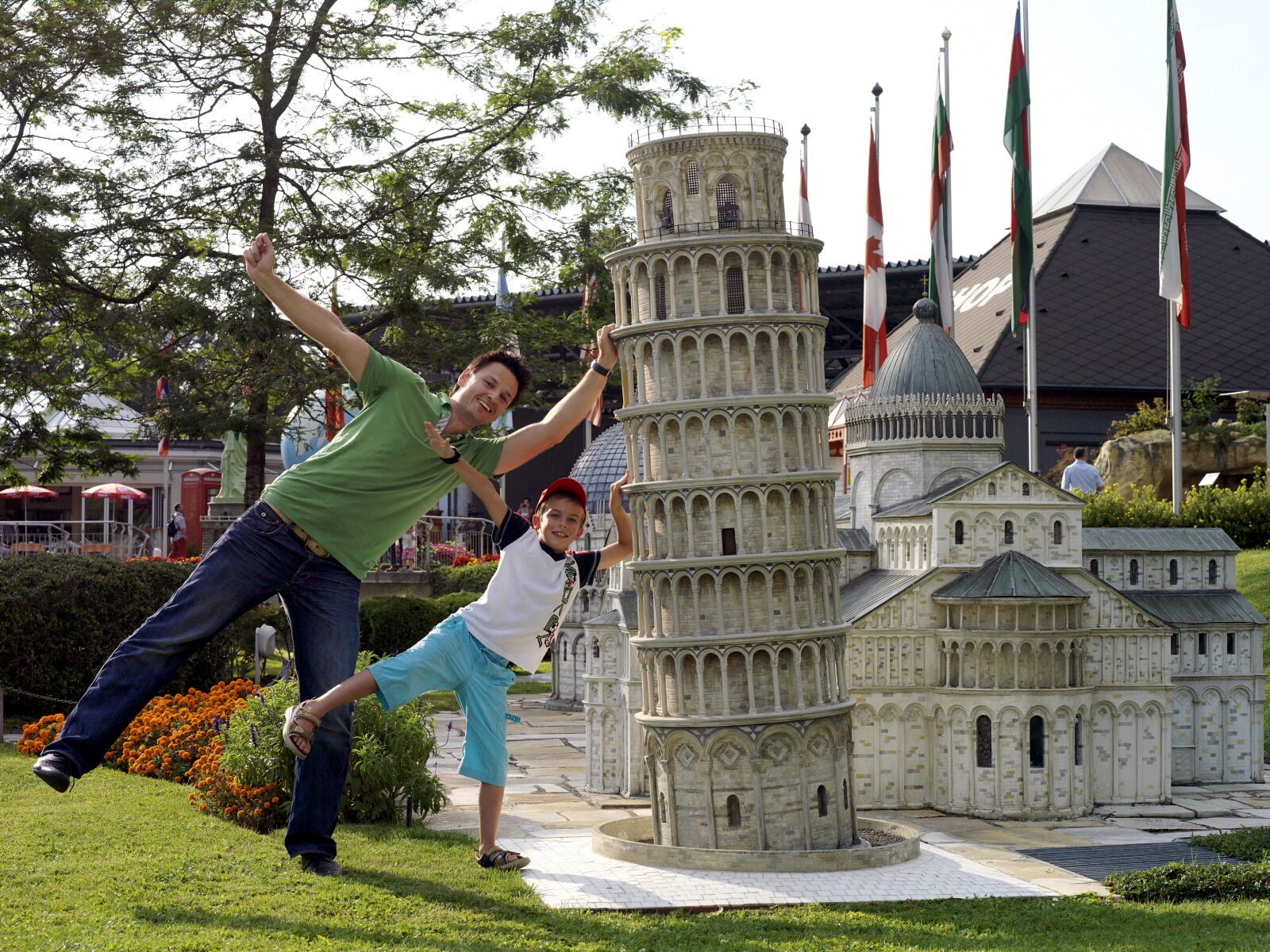 A model of the Leaning Tower of Pisa surrounded by flags, with a person and a child posing in front.