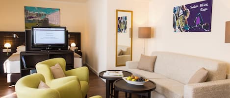 A modern living room with a sofa, two armchairs, a coffee table with fruit, a TV, and artwork on the walls.