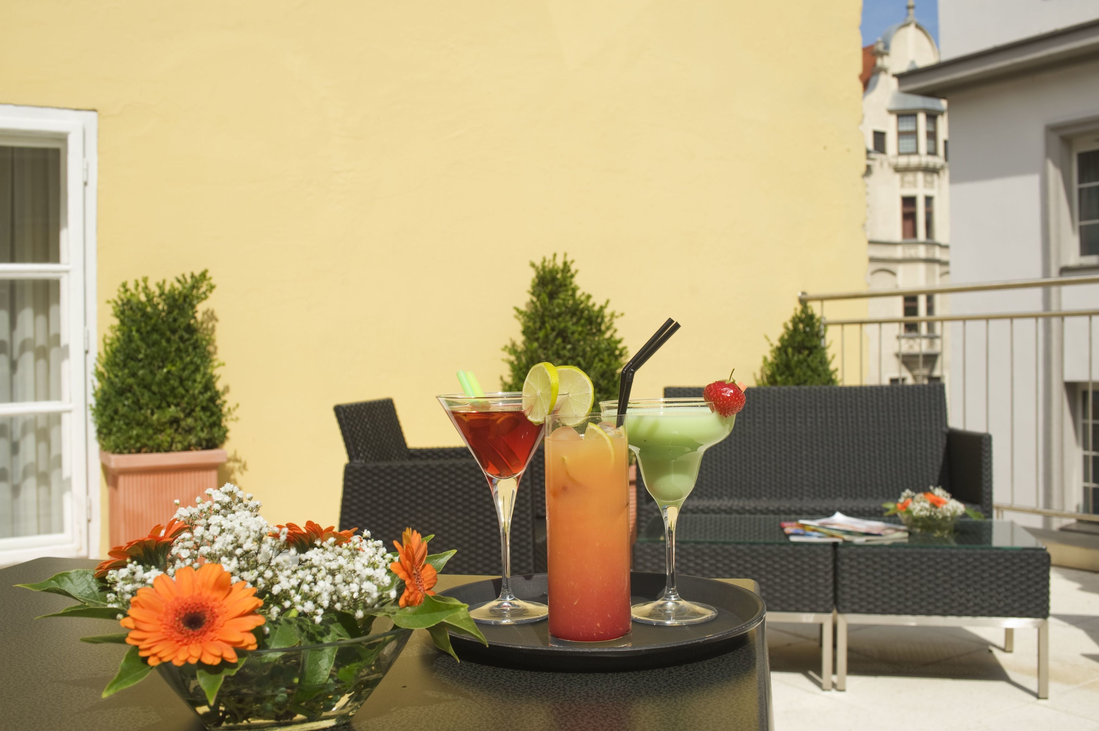 Ein Tisch mit drei Getränken, einer Blumenkomposition und Gartenmöbeln vor einer gelben Wand.