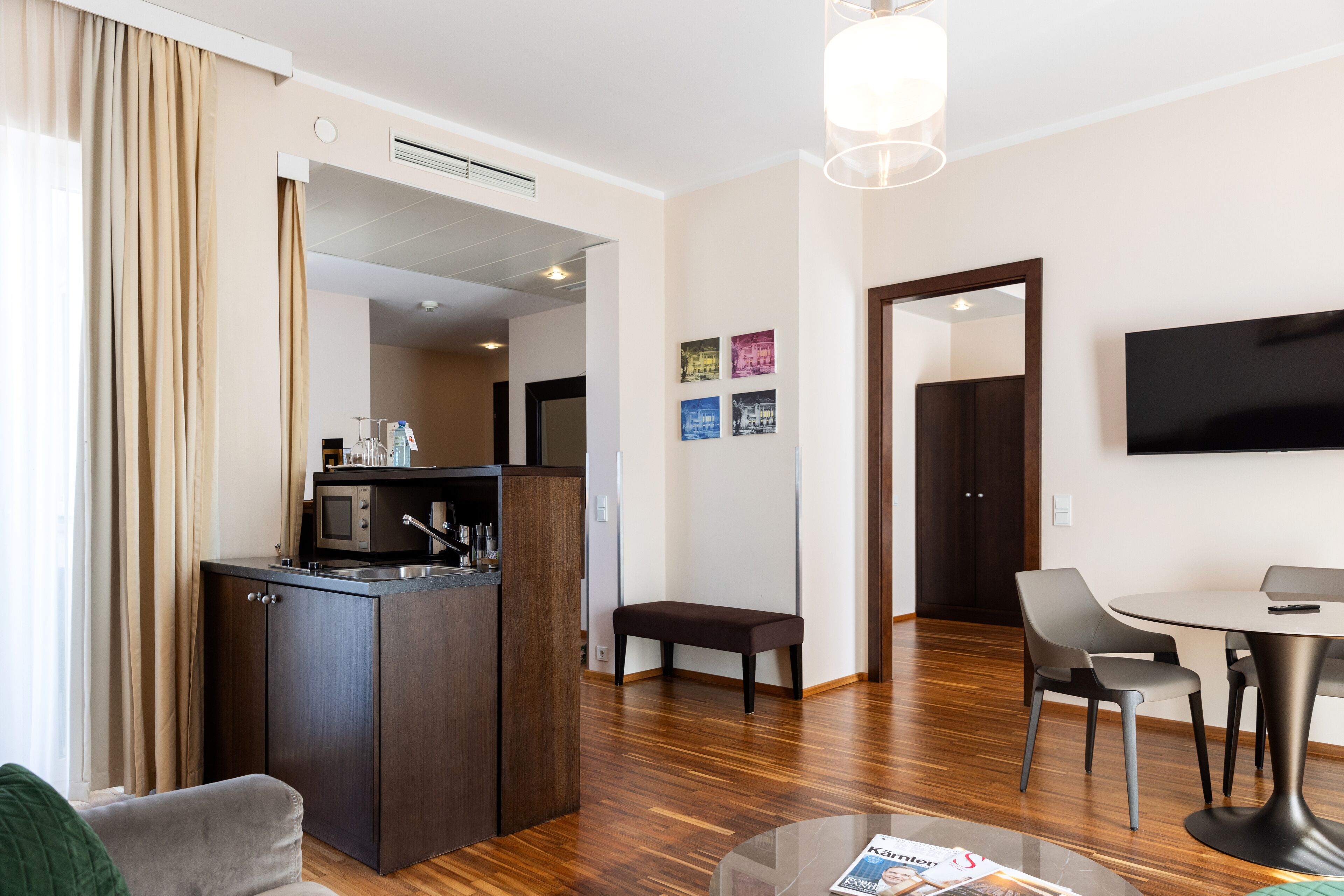 Una habitación de hotel moderna con kitchenette, una zona de comedor con mesa y sillas, un banco y un televisor de pantalla plana montado en la pared.