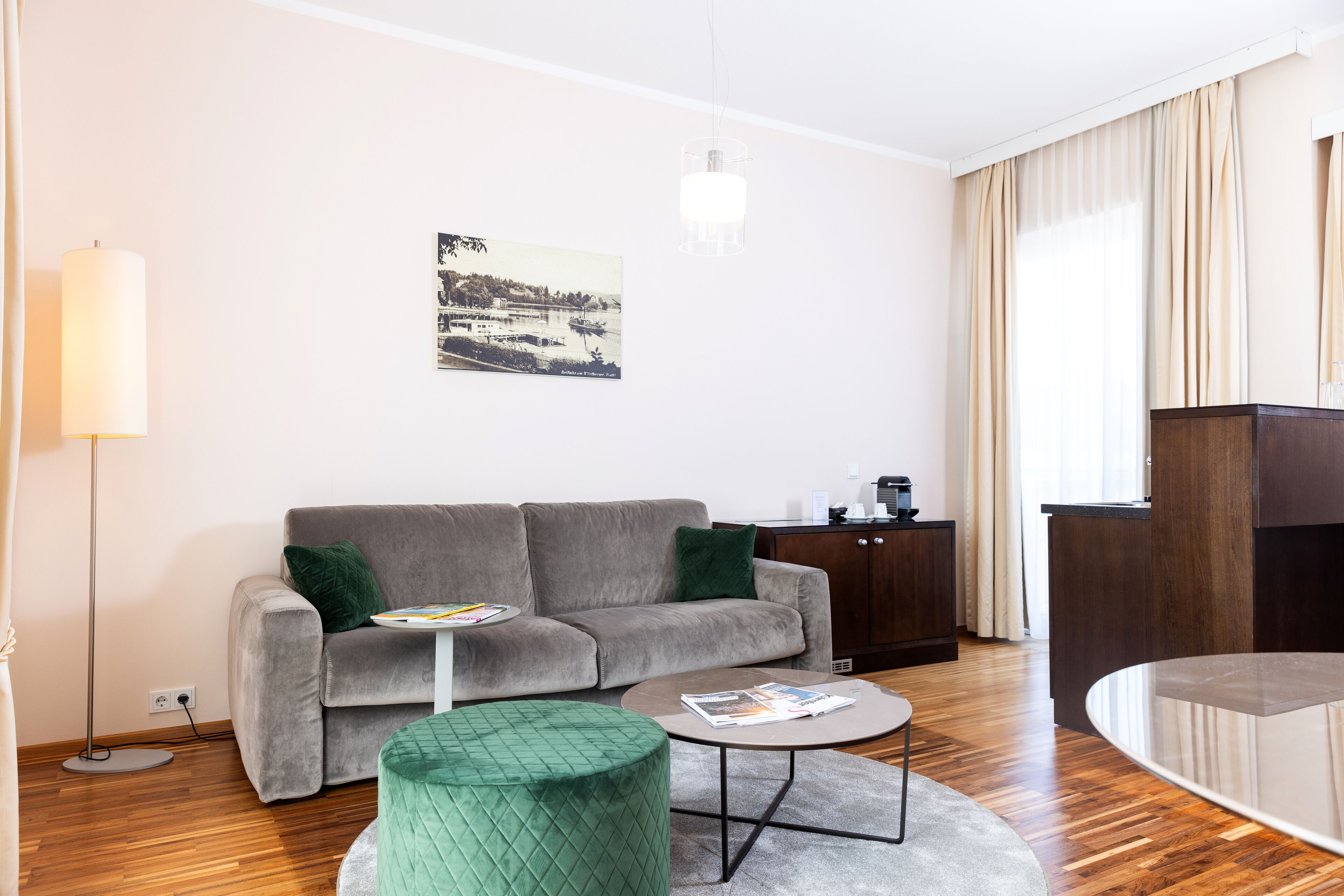 Una sala de estar moderna con un sofá gris, una mesa redonda de centro, un otomano verde, una lámpara de pie y un cuadro en la pared.