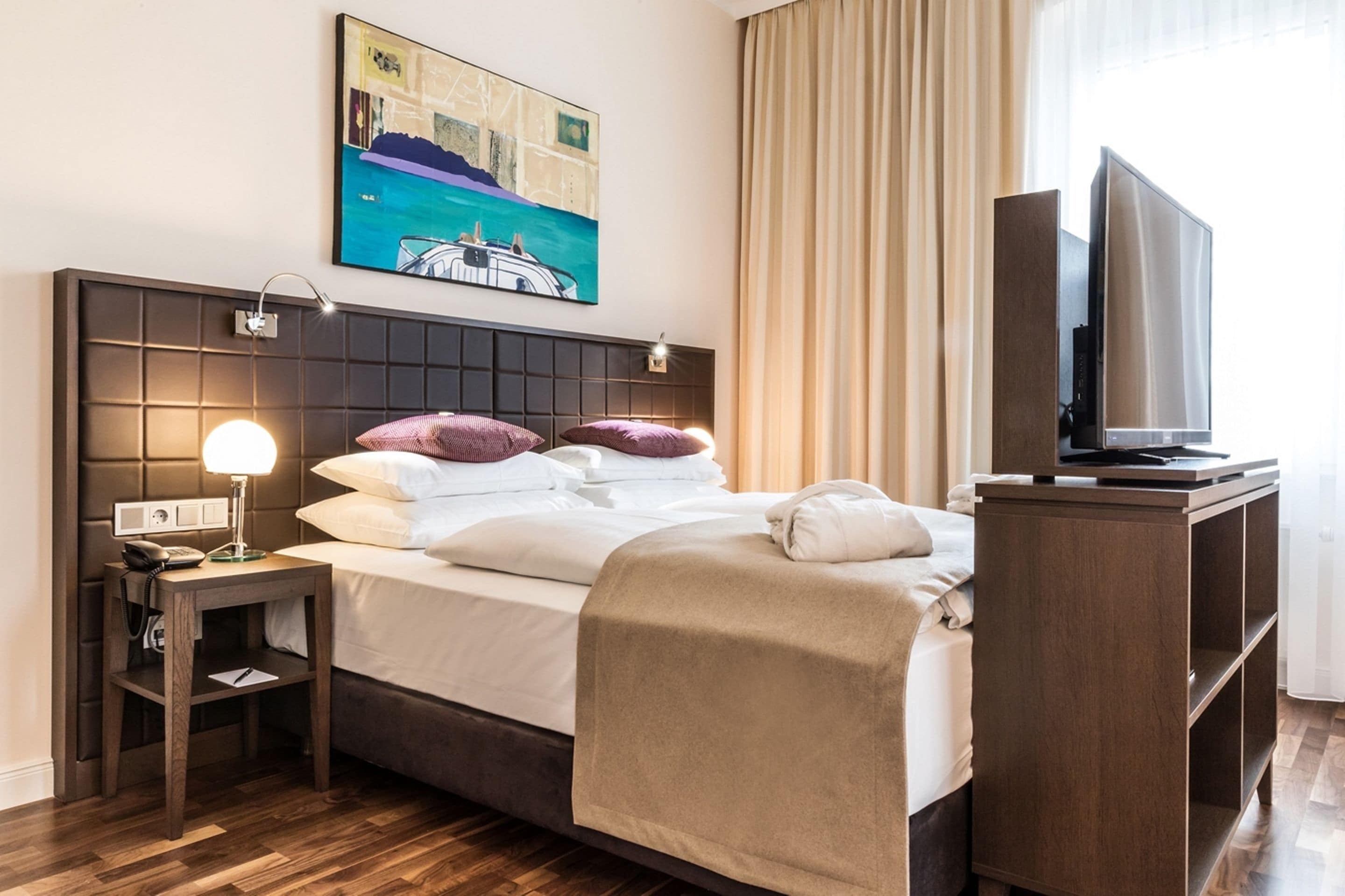 A hotel room with a bed, a bedside table with a lamp, a TV, and a painting on the wall.