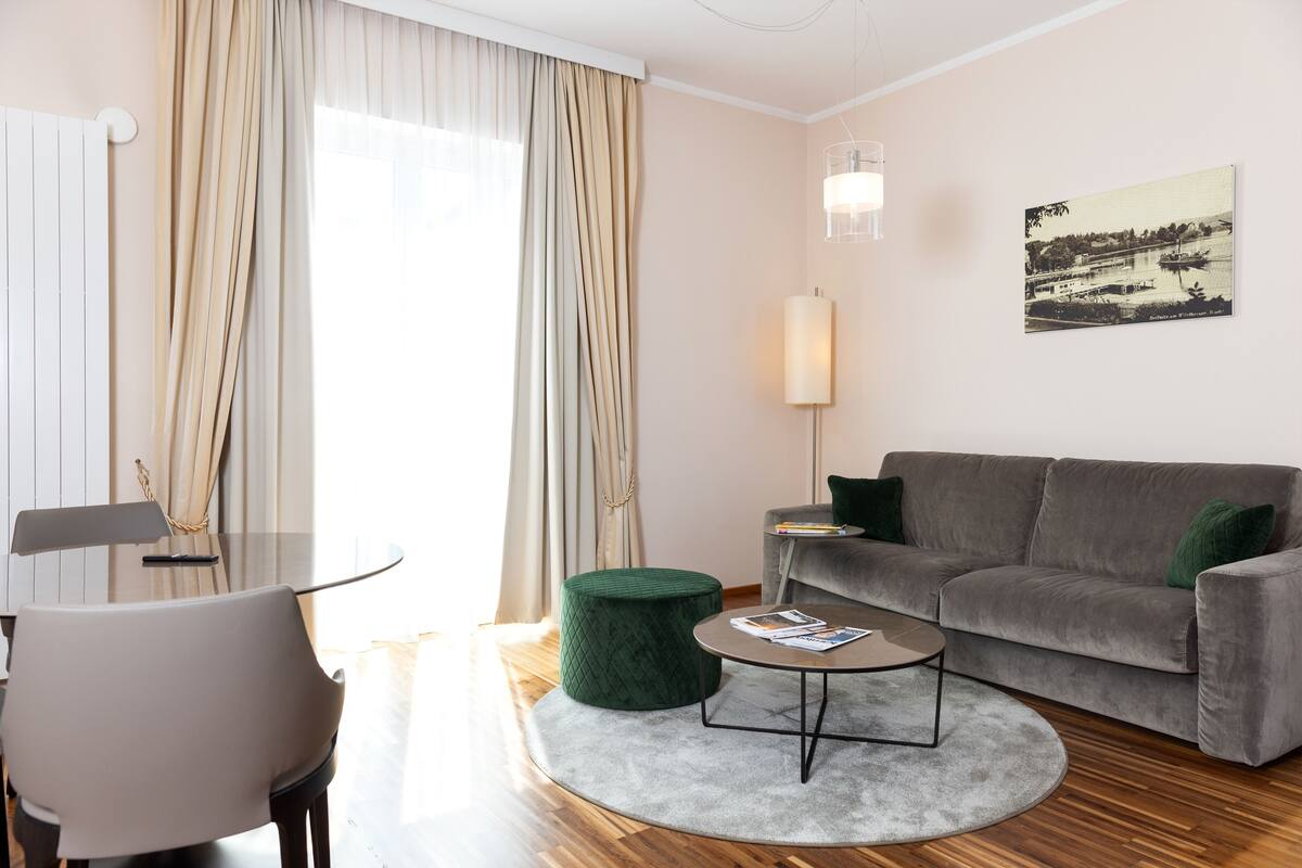 A modern living room with a grey sofa, a round coffee table, a green ottoman, a dining table with chairs, and a large window with curtains.