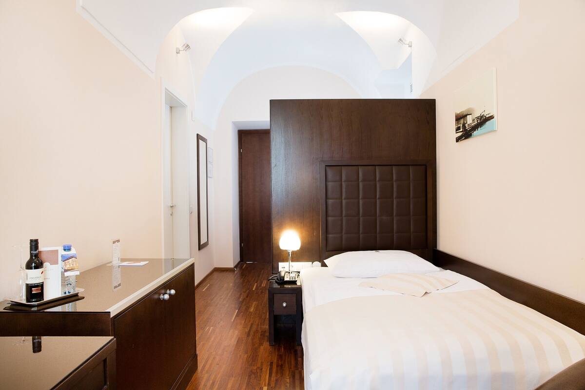 A hotel room with a wooden headboard, a bedside table with a lamp, a bed with white bedding, and a wooden dresser.