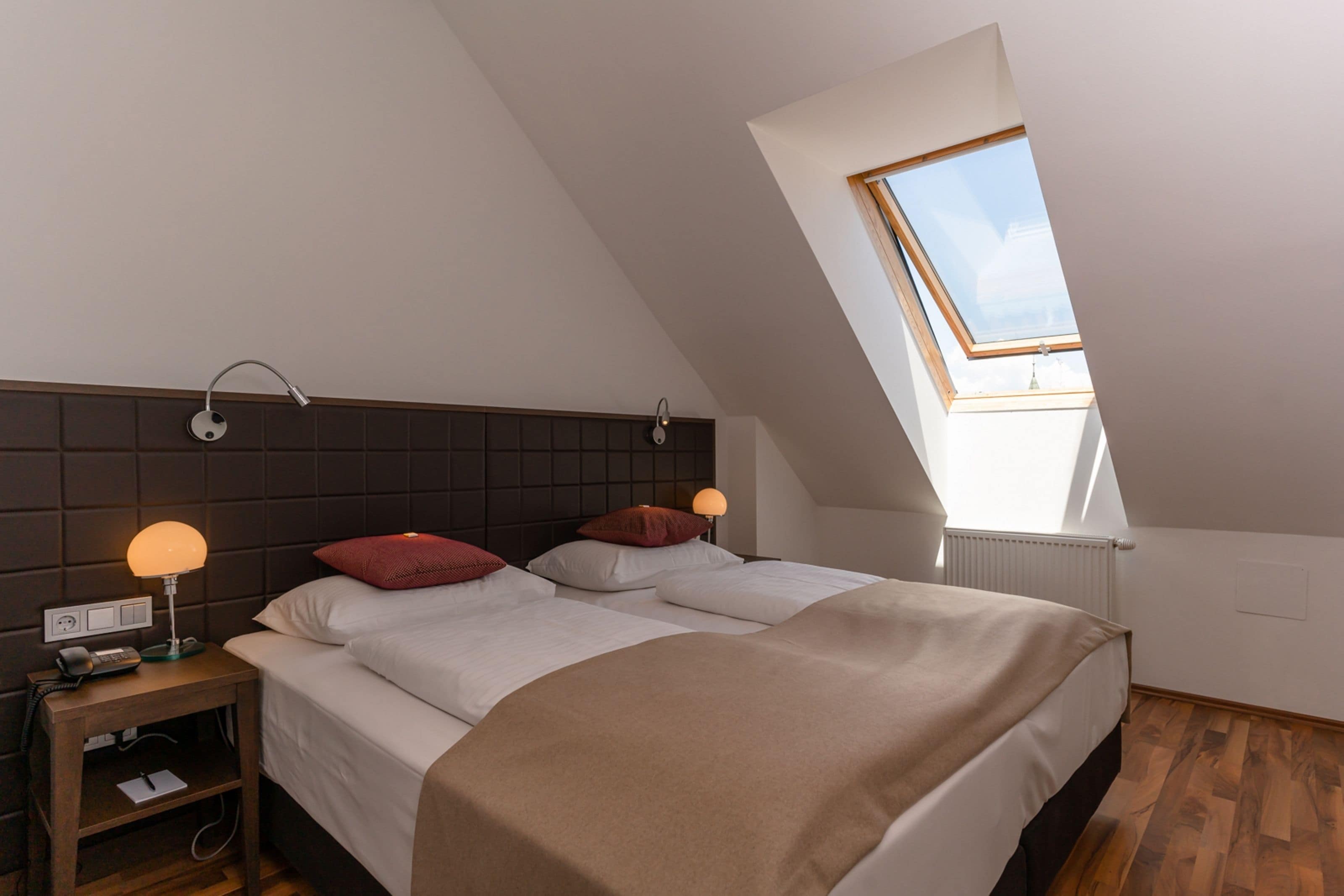 A bedroom with two beds, a nightstand with a lamp, a window, and a radiator.