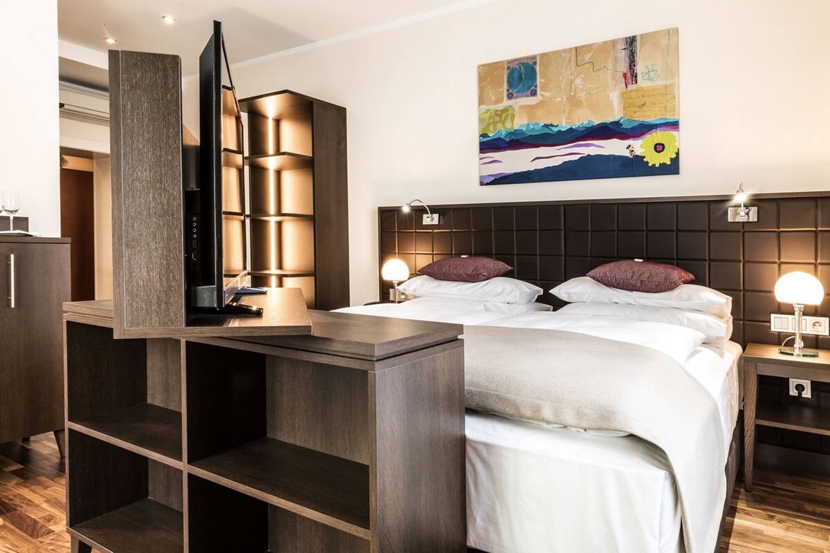A modern hotel room with a bed, bedside tables, a TV, and a painting on the wall.