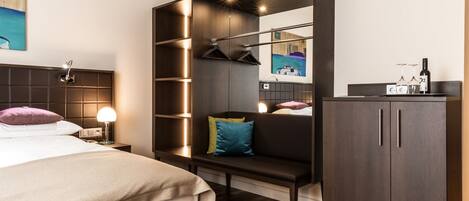 A modern hotel room with a bed, a bench, a wardrobe, and a small table with a bottle and glasses.