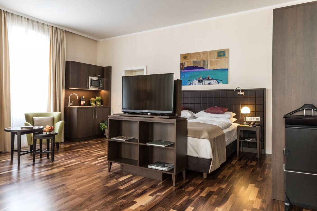 A modern hotel room with a bed, a TV, a desk, a chair, a small kitchenette, and a painting on the wall.