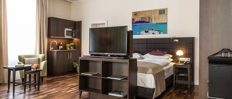 Een moderne hotelkamer met een bed, een televisie, een bureau, een stoel, een kleine kitchenette en een schilderij aan de muur.