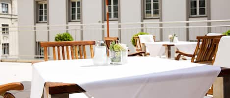 Mesa colocada al aire libre con mantel blanco, sillas de madera y un florero de vidrio con flores.