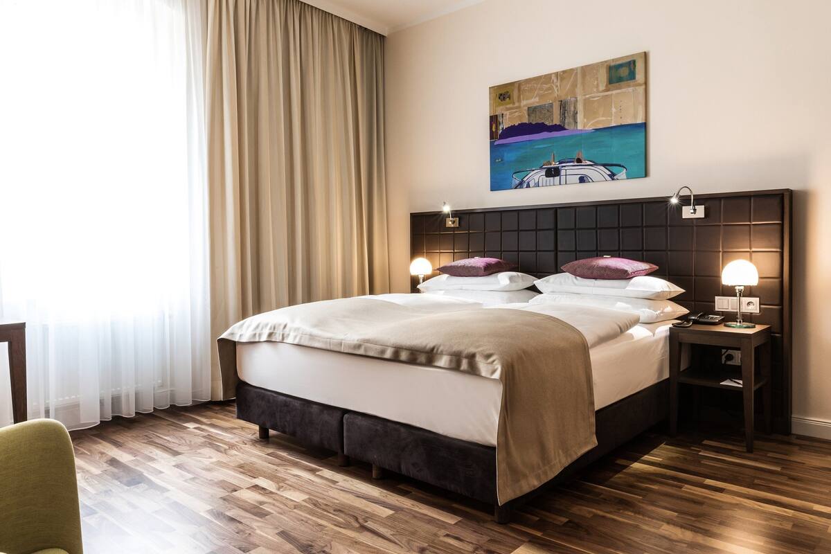 A hotel room with a large bed, a headboard with a light fixture, a bedside table with a lamp, and a painting on the wall.