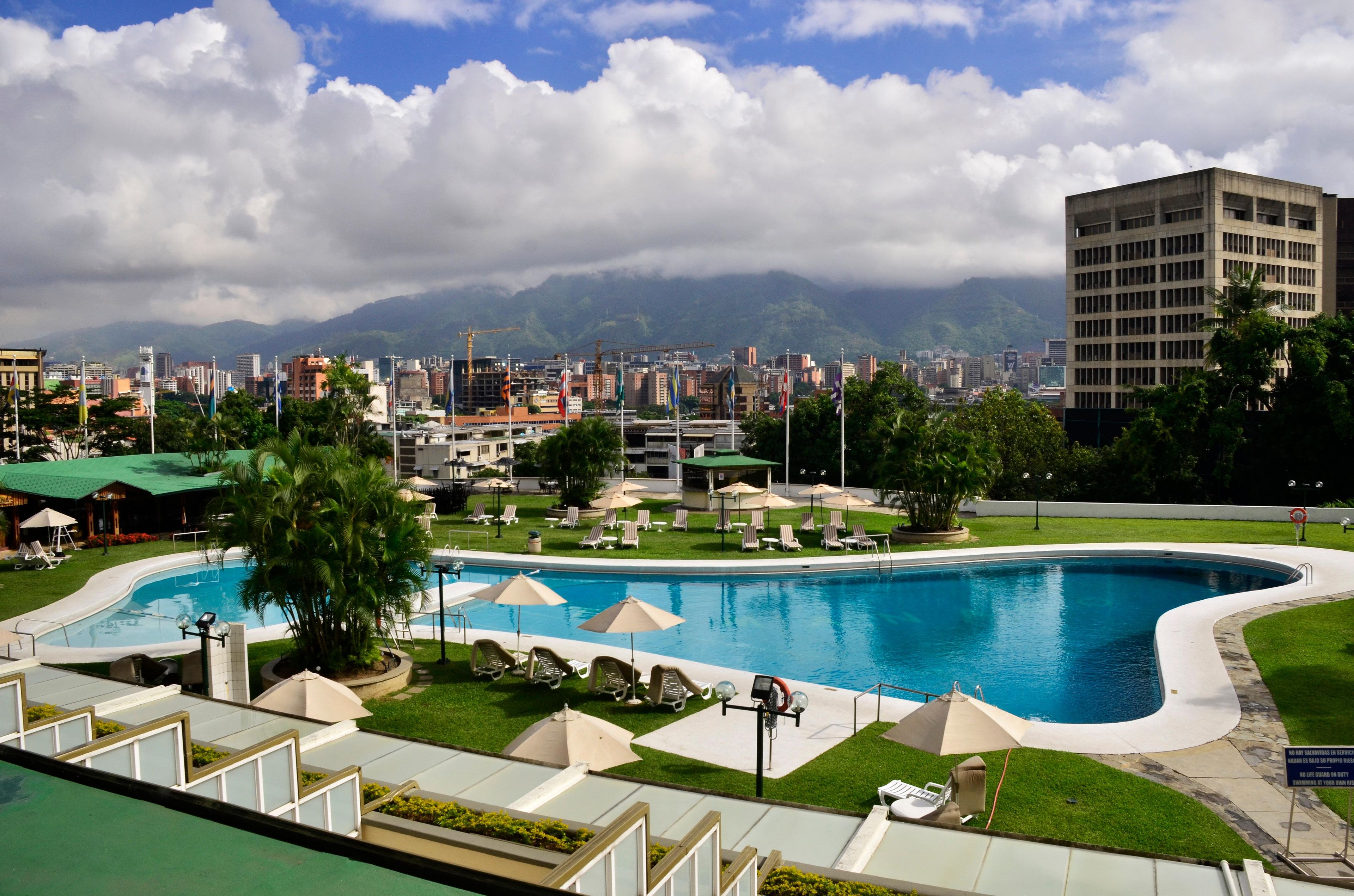 A swimming pool with lounge chairs and umbrellas, surrounded by greenery and a cityscape with mountains in the background.